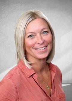 Smiling woman with straight blonde hair in a chin-length bob, wearing a coral button-up shirt and a gold necklace, posed against a light gray studio backdrop.