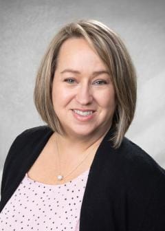 Smiling woman with straight shoulder-length blonde hair, wearing a pink polka dot blouse with a black cardigan and a small pendant necklace, posed against a light gray studio backdrop.