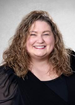 Smiling woman with long curly light brown hair, wearing a black top with sheer sleeves, posed against a light gray studio backdrop.