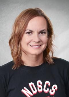 Smiling woman with shoulder-length light brown hair, wearing a black NDSCS shirt, posed in front of a gray studio background