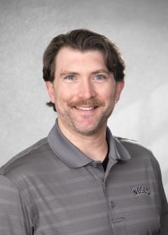 Smiling man with wavy brown hair and mustache, wearing a gray NDSCS polo, posed in front of a gray studio background