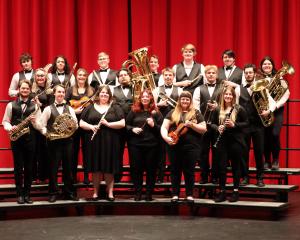 students standing on stage in formal concert attire, holding various instruments