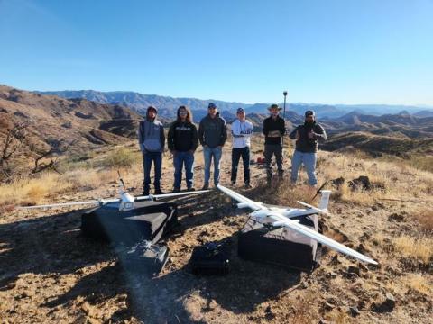 Six students stand around two large drones on the ground. The setting is the desert landscape of Arizona.