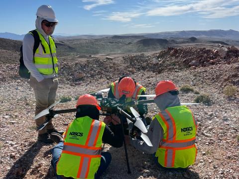 3 students sit by a large drone, working on it. Another student stands nearby, watching them. All are wearing high-visibility vests and hard hats.